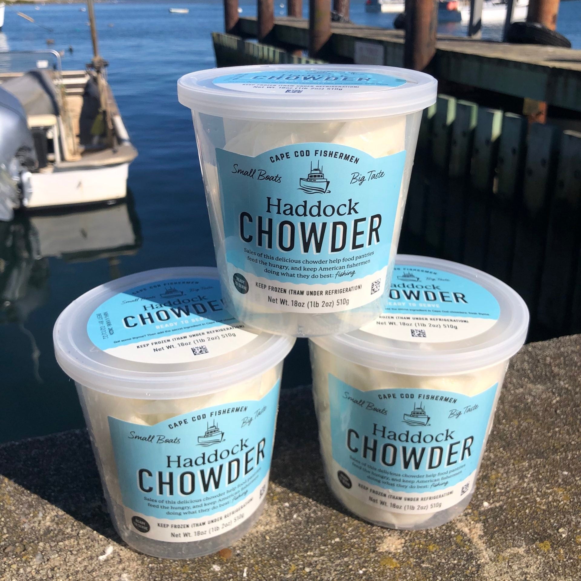 Three containers of Haddock Chowder by Cape Cod Fisherman on a dock with water and boats in the background.